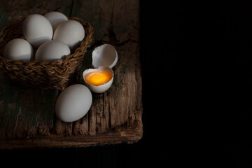 On an old table, white eggs in a wicker basket, other eggs around it, one of them split in two, with its yolk illuminated by a dim ray of light, it is a beauty. All in dark background
