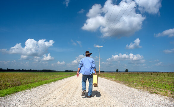 Man walking down one lane country dirt road wearing cowboy hat, carrying old retro suitcase, blue sky, clouds, travel, traveling, independent, freedom, 