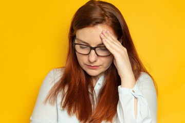 Unhappy young woman with red long hair and glasses suffers a headache, migraine on a yellow wall...