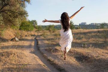 happy girl runs in the field along the path, bouncing and waving her arms. Against the background of the summer setting sun