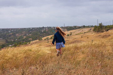 Adorable little girl in denim jacket, blue plaid dress running in yellow wild grass field. Happy stylish long blonde hair child on countryside landscape. Cute kid walking outdoor rural road trip.