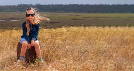 Little blonde girl in denim jacket, black sunglasses, blue dress with vintage suitcase grass field banner. Stylish hitchhiker child with long hair countryside landscape trip. Kid walking outdoor road