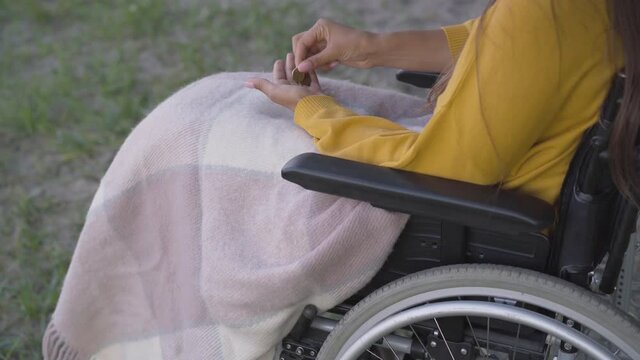 Unrecognizable young woman in wheelchair counting coins outdoors. Poor paralyzed Caucasian invalid having little money. Social problems of disabled people.