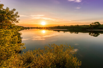 Naklejka na ściany i meble Scenic view at beautiful summer river sunset with reflection on water with green bushes, grass, golden sun rays, calm water ,deep blue cloudy sky and glow on a background, spring evening landscape