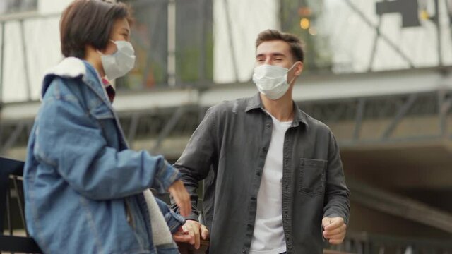 Medium shot of Caucasian young man talking with Asian girlfriend leaning on balcony railing, both wearing medical face masks