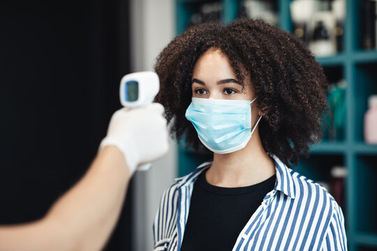 Young black woman in protective mask getting temperature check up in salon