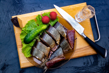 a shot of vodka stands next to a plate of cucumber, radish, and chopped smoked herring