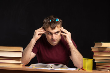 a young student sits with his hands behind his head at a table with books on a black wall background. office worker examines documents. European look. distance learning. preparation for exams.