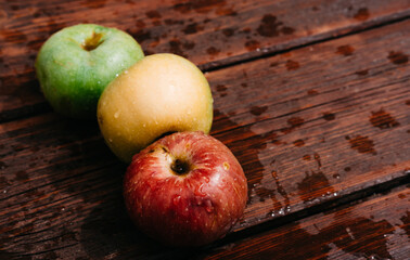 Fresh apples, autumn harvest. Three apples - green, yellow, and red-lie on a wooden table. Fresh apples on a wood textured brown background, mahogany material.
