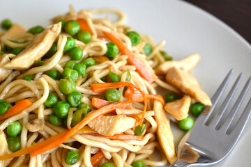 Chicken chow mein on a white plate with a fork on the right side.  Stir fry noodles with chicken, carrot and green peas.  