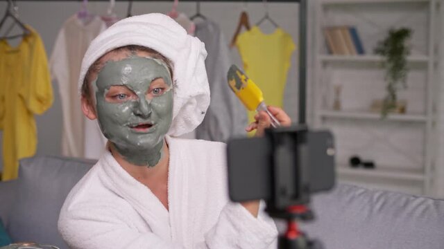 A Girl With a White Towel Put on Her Face a Green Mask During Blogging.Teenager Girl Vlogging About Сosmetic Сare. Girl Shooting Beauty Videoblog.Girl in a White Coat Talking With Followers