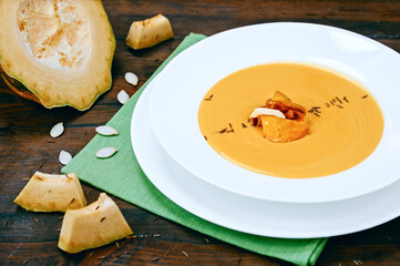 Portion of creamy pumpkin soup puree with fresh vegetable, seeds and smoked ham in white plate