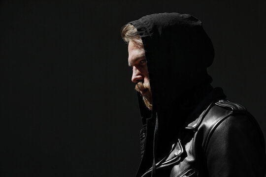 Brutal man with ginger thick beard, looks thoughtfully aside, wears hoodie posing over black background with free space.