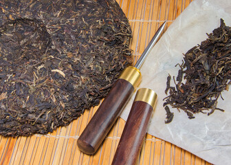 Pu Erh Teefladen auf Bambusmatte mit Teepicker und losem Tee auf weißem Papier