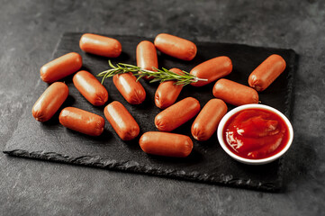 raw mini sausages with spices and ketchup on a slate board on a stone background