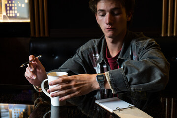 a young man is drinking tea at the table. a person comes up with a plan or idea before writing it down in a notebook.
