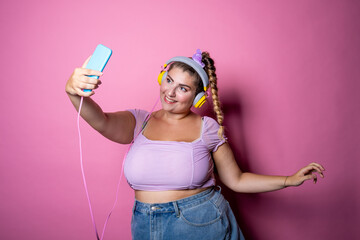 Young beautiful plus size woman taking selfie on pink background - Isolated chubby young woman live streaming wearing headphones studio shot