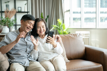 Social distancing or new normal concept.Senior asian couple sitting on sofa in home playing smartphone and video conference with grandchild.They are smiling to spend their time together with happiness