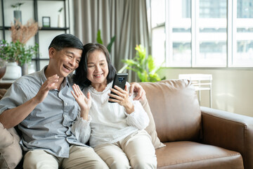 Fototapeta premium Social distancing or new normal concept.Senior asian couple sitting on sofa in home playing smartphone and video conference with grandchild.They are smiling to spend their time together with happiness