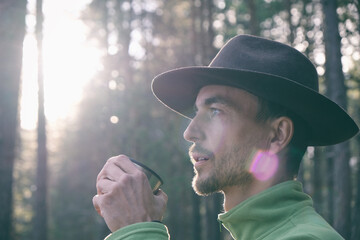 Portrait of bearded millennial man in felt hat drinking hot tea in woods on sunset. Tourist...
