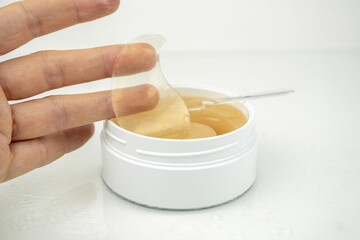 take golden under eyes patches with special spoon from a white container, anti aging skin care, white wet background
