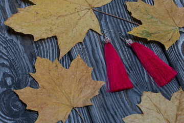 Tassel earrings made of silk threads. Nearby are dried maple leaves. They lie on pine boards painted in black and white. Autumn gifts for loved ones.