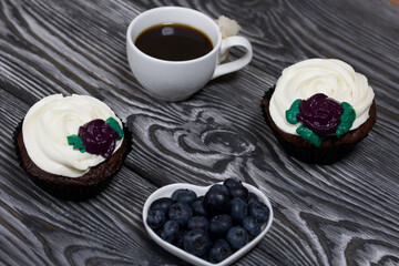 Chocolate muffins with cream flower. A cup of black coffee. Saucer with blueberries. Lies on pine boards painted black and white.