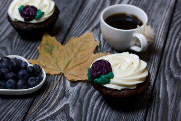Chocolate muffins with cream flower. A cup of black coffee. Saucer with blueberries. Dried maple leaf. Lies on pine boards painted black and white.