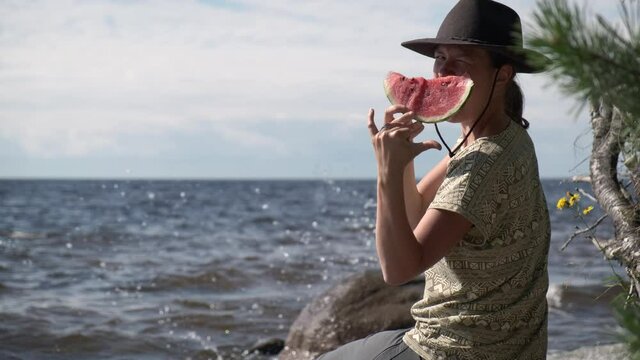 Hipster woman in felt hat sitting near sea and eating fresh watermelon, showing by hand surfing sign shaka aloha. Summer vacation.