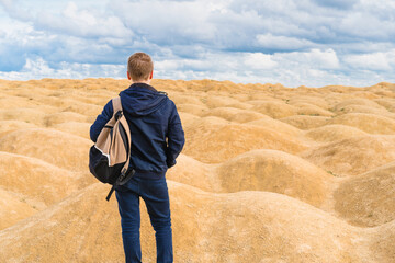 Sporty blond man with a backpack stands with his back to the camera in the desert with sand dunes of bizarre shape