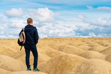 Sporty blond man with a backpack stands with his back to the camera in the desert with sand dunes of bizarre shape