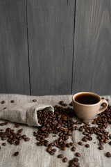 Coffee scattered from a linen bag and a cup of black coffee Espresso or Americano standing on a wooden table. Fresh arabica coffee beans on linen textile. Aromatic freshly brewed coffee.