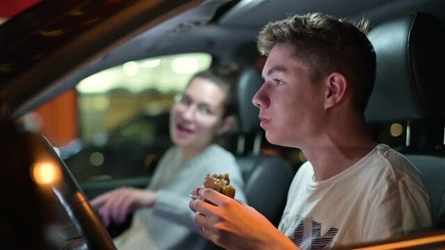Young guy, teenager, eating a burger in the car in the evening in the parking lot, next to him sits a young woman