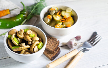 Salted mushrooms and pickled cucumbers in bowls on a white wooden table