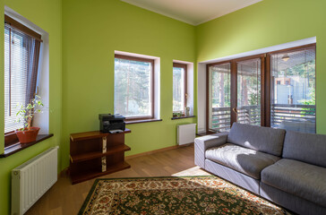 Modern interior of home office room with green walls in luxury private house. Cozy couch. Printer on wooden shelf. Glass door to terrace.