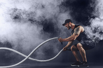Bearded athletic looking bodybulder work out with battle rope on dark studio background with smoke....