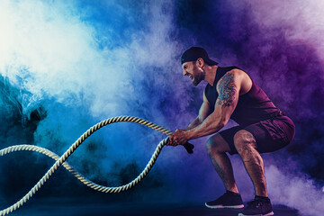 Bearded athletic looking bodybulder work out with battle rope on dark studio background with smoke. Strength and motivation.