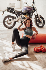 Sport woman resting after training and sitting with gloves and bag on floor and modern motorbike on the background.