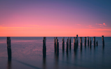 Obraz premium Tranquil seascape with ruined pilings of the pier in the seawater. Warm glowing pink sunrise over blue water. Long exposure image.