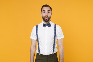 Shocked amazed young bearded man 20s wearing white shirt bow-tie suspender posing keeping mouth open saying wow looking camera isolated on bright yellow color wall background, studio portrait.