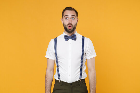 Shocked amazed young bearded man 20s wearing white shirt bow-tie suspender posing keeping mouth open saying wow looking camera isolated on bright yellow color wall background, studio portrait.