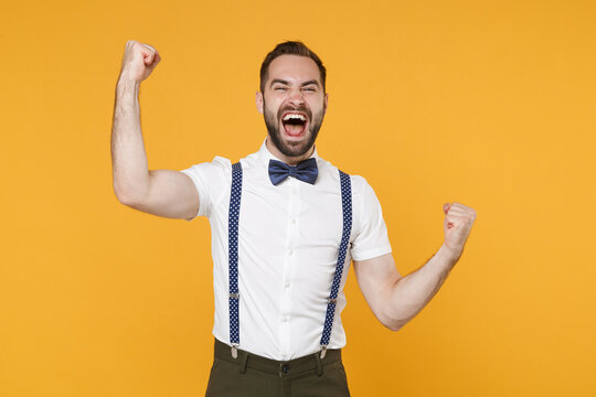 Overjoyed screaming young bearded man 20s wearing white shirt bow-tie suspender posing clenching fists doing winner gesture say yes isolated on bright yellow color wall background, studio portrait.