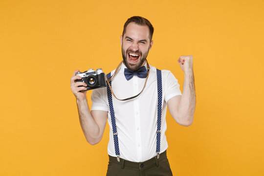 Happy young bearded man 20s wearing white shirt bow-tie suspender posing taking picture with retro vintage photo camera doing winner gesture isolated on bright yellow color background studio portrait.