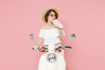 Pretty young brunette woman 20s wearing white summer clothes hat eyeglasses hold in hand plastic cup of cola or soda sitting driving moped isolated on pastel pink colour background studio portrait.