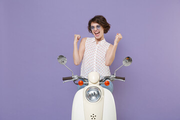 Happy joyful young woman 20s in white dotted shirt glasses clenching fists doing winner gesture keeping eyes closed sitting driving moped isolated on pastel violet colour background, studio portrait.