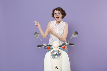 Shocked surprised young brunette woman in white dotted shirt glasses point index fingers aside on mock up copy space sitting driving moped isolated on pastel violet colour background studio portrait.