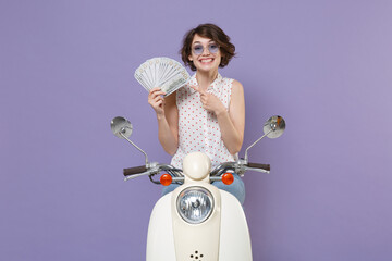 Funny young brunette woman 20s in white dotted shirt glasses pointing index finger on fan of cash money in dollar banknotes driving moped isolated on pastel violet colour background studio portrait.