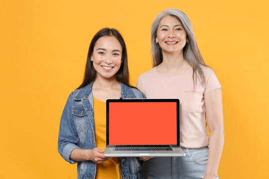 Smiling family asian women girls gray-haired mother brunette daughter in casual clothes hold laptop computer with blank empty screen mock up copy space isolated on yellow background studio portrait.