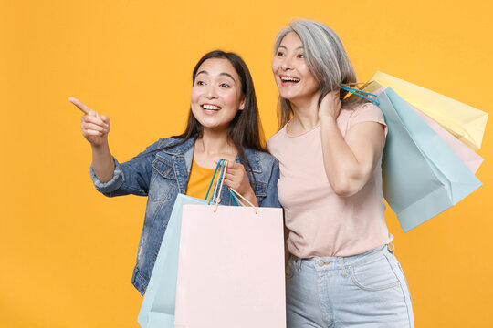 Excited family asian female women girls mother and daughter in casual clothes hold package bag with purchases after shopping pointing index finger aside isolated on yellow background studio portrait.