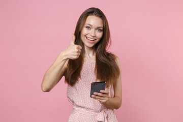 Cheerful young brunette woman 20s wearing pink summer dotted dress posing using mobile cell phone typing sms message showing thumb up isolated on pastel pink color wall background studio portrait.